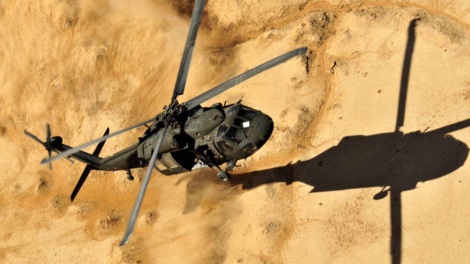 A Sikorsky Black Hawk powered by T700 landing on a sandy desert terrain, casting a large shadow on the ground below. The helicopter's rotors are spinning, and dust is being stirred up by its movement.