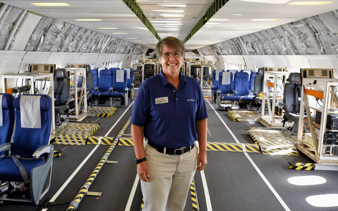 A person wearing a blue shirt and khaki pants stands inside a spacious aircraft cabin equipped for testing. The interior features rows of blue seats, exposed wiring, and specialized equipment along the walls and floor. Yellow and black caution tape marks sections of the floor, emphasizing the experimental setup. The person is smiling and positioned at the center of the image.