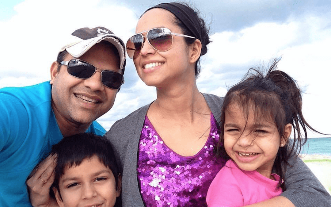A family of four poses together on a beach, with the ocean and cloudy sky visible in the background.