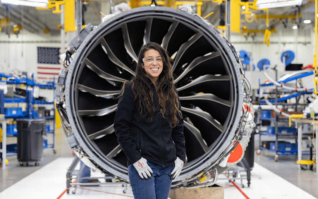 A person with long hair stands in front of a large aircraft engine in an industrial setting. They are wearing a black jacket and white gloves, smiling at the camera. The background includes various machinery, equipment, and workstations, indicating a manufacturing environment.