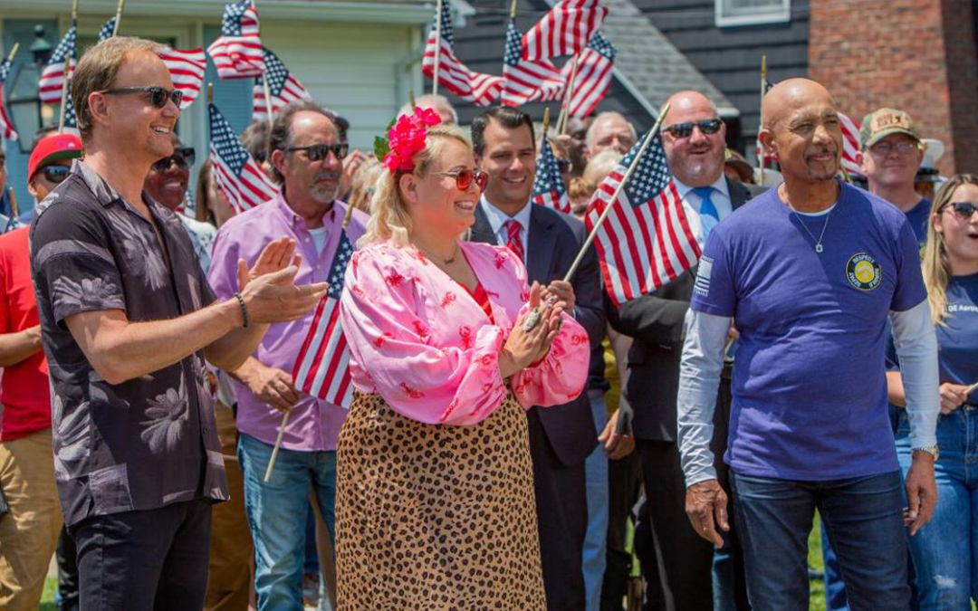 Image of Jennifer Bertram and Montel Williams standing with a crowd all smiling and holding American flags. 