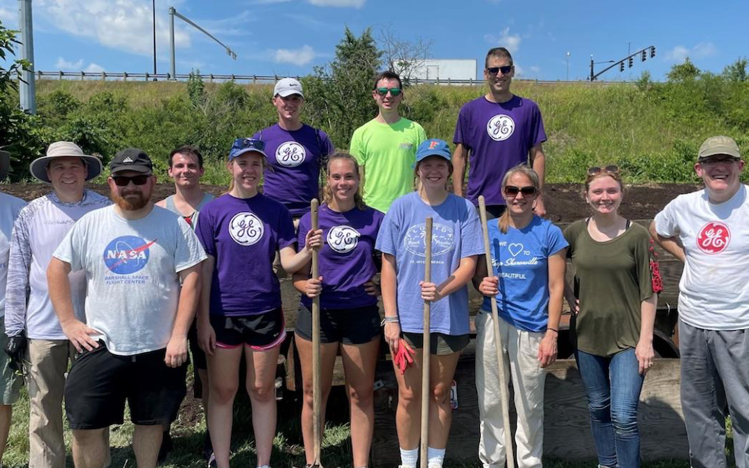 A group of GE employees and volunteers standing outdoors, holding gardening tools and wearing various GE and NASA t-shirts.